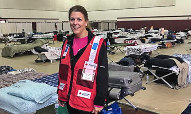A female woman wearing a Red Cross vest stands among cots in a gymnasium being used as a temporary shelter.