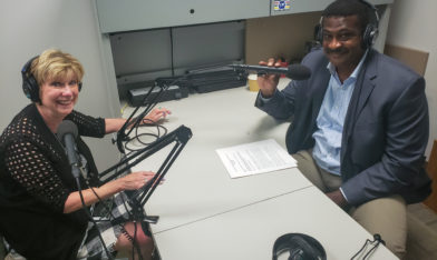 photo showing Mary Jo Monahan and Angelo McClain sitting at a desk with microphones in a recording studio.