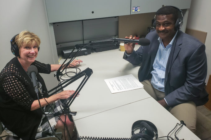 photo showing Mary Jo Monahan and Angelo McClain sitting at a desk with microphones in a recording studio.